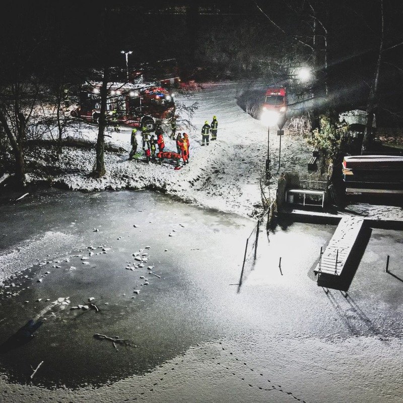 Eisrettung bei Nacht an einem zugefrorenen See, Einsatzkräfte und Feuerwehrfahrzeuge am verschneiten Ufer im Licht von BASELIGHT
